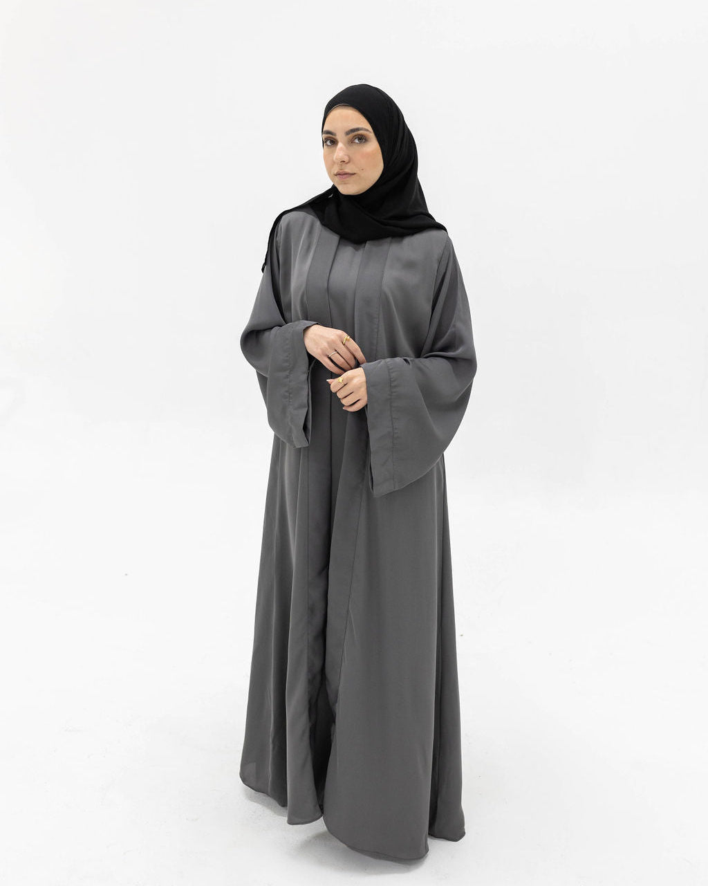 A woman wearing a long, flowing dark gray abaya with wide sleeves and a black hijab, standing against a plain white studio background.