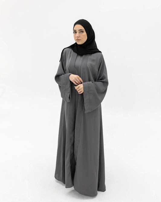 A woman wearing a long, flowing dark gray abaya with wide sleeves and a black hijab, standing against a plain white studio background.